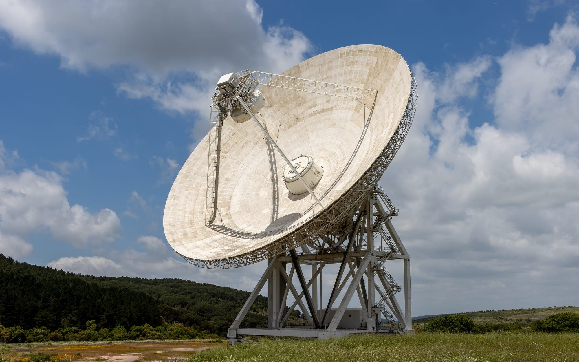 Large white parabolic radio telescope dish on a steel mount among green hills under clouds
