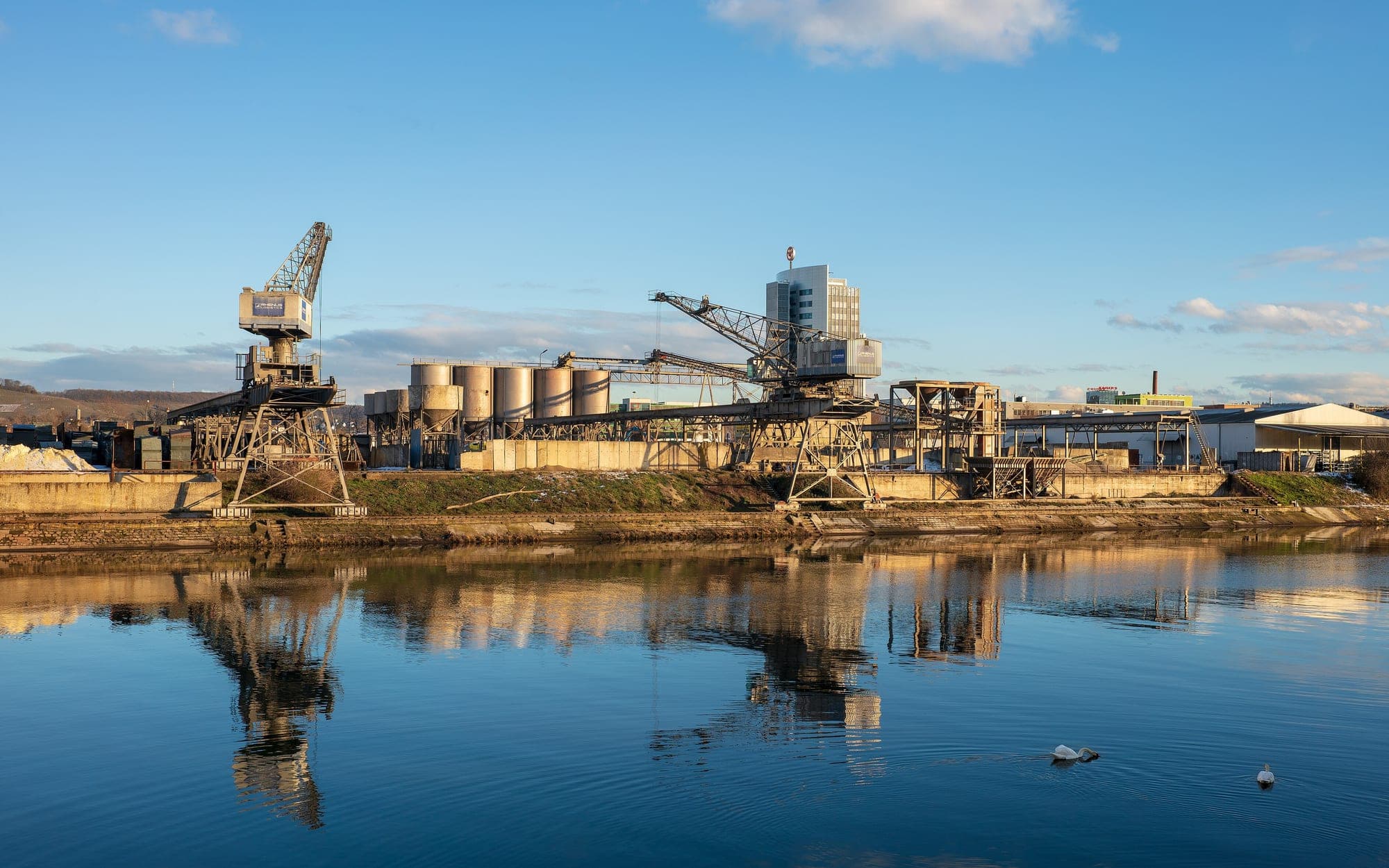 Port cranes, silos and conveyors on a riverbank reflected in calm water in the evening