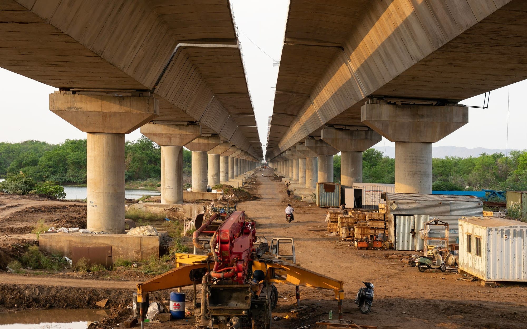 Concrete piers and two parallel spans of a road bridge under construction, with a building site and machinery below