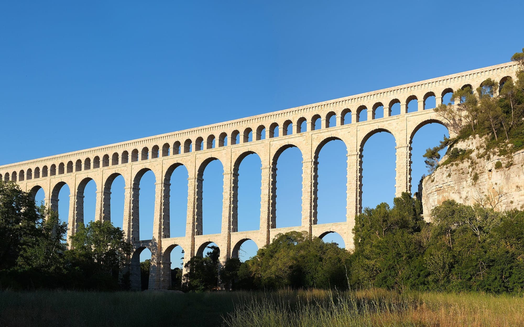 Multi-tiered stone arches of the Roquefavour aqueduct against a blue sky above green trees