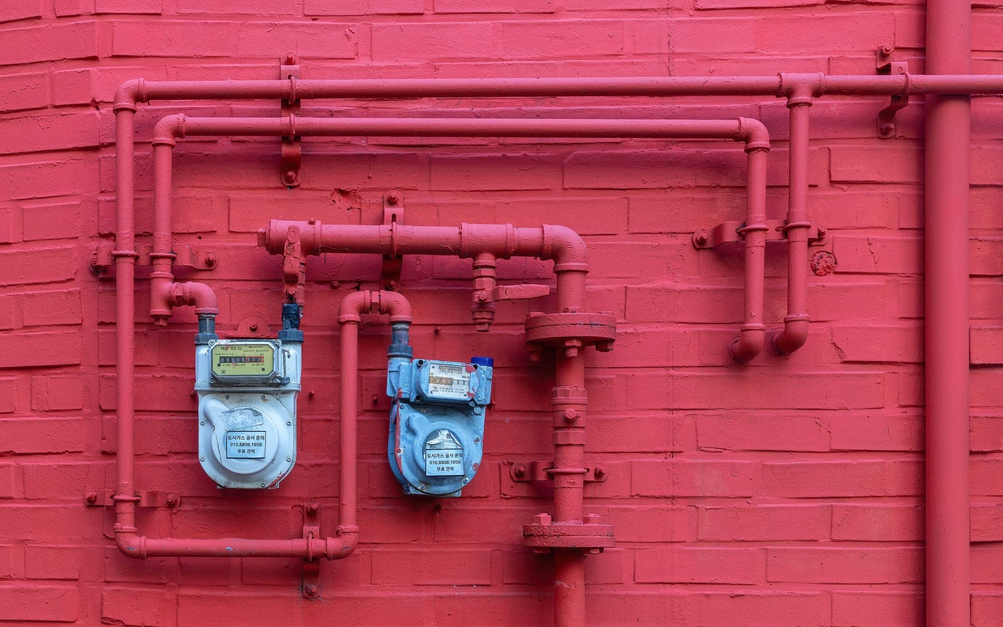 Two blue meters on red pipes running across a matte red painted brick wall
